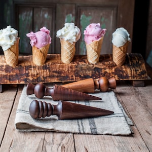 Pode incluir: Uma variedade de cones de sorvete com sorvete de baunilha e morango, exibidos em um suporte de madeira. Abaixo, três moldes de madeira para cones de sorvete repousam sobre um pano de linho, em um fundo de madeira rústica.