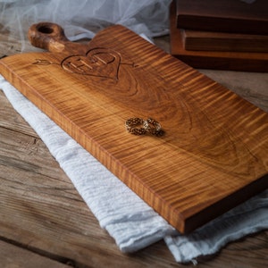 Peut inclure: Une planche à découper en bois avec un cœur gravé et deux anneaux dorés dessus. La planche a une finition en bois naturel et un motif de grain unique.
