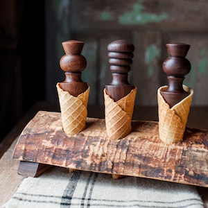 Può includere: Tre porta coni gelato in legno scuro con forme uniche, ciascuno in un cono a cialda. I porta coni sono esposti su una tavola di legno rustica, con uno sfondo sfocato.