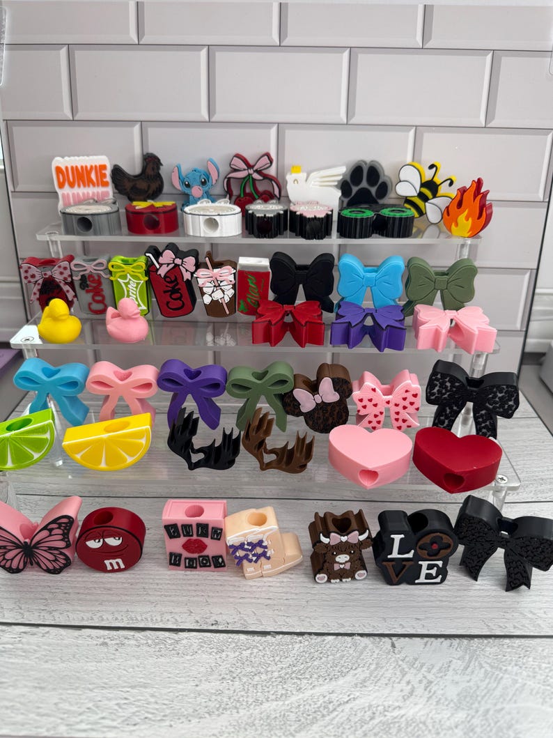 May include: A collection of decorative pencil sharpeners in various shapes and colors, including bows, hearts, and cartoon characters. Some sharpeners feature text like "Dunkie" and "Love". The sharpeners are displayed on clear acrylic shelves.
