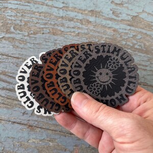 May include: A set of six round leather patches in various shades of brown, gray, and white. Each patch features the text "Overstimulated" in a circular design with a stylized flower and smiley face.