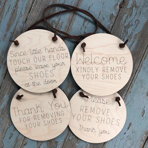 Puede incluir: Cuatro letreros redondos de madera con correas de cuero marrón que cuelgan de ellos. Los letreros tienen diferentes mensajes sobre quitarse los zapatos. El primer letrero dice "Since little hands touch our floor please leave your shoes at the door". El segundo letrero dice "Welcome kindly remove your shoes". El tercer letrero dice "Please remove your shoes thank you". El cuarto letrero dice "Thank you for removing your shoes".
