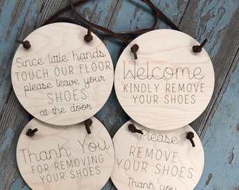 May include: Four round wooden signs with brown leather straps hanging from them. The signs have different messages about removing shoes. The first sign says "Since little hands touch our floor please leave your shoes at the door". The second sign says "Welcome kindly remove your shoes". The third sign says "Please remove your shoes thank you". The fourth sign says "Thank you for removing your shoes".