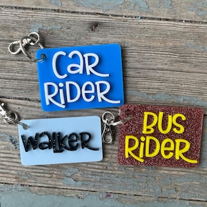 May include: Three keychains with different colored backgrounds and text. The first keychain is blue with white text that says "car rider". The second keychain is white with black text that says "walker". The third keychain is brown with yellow text that says "bus rider".
