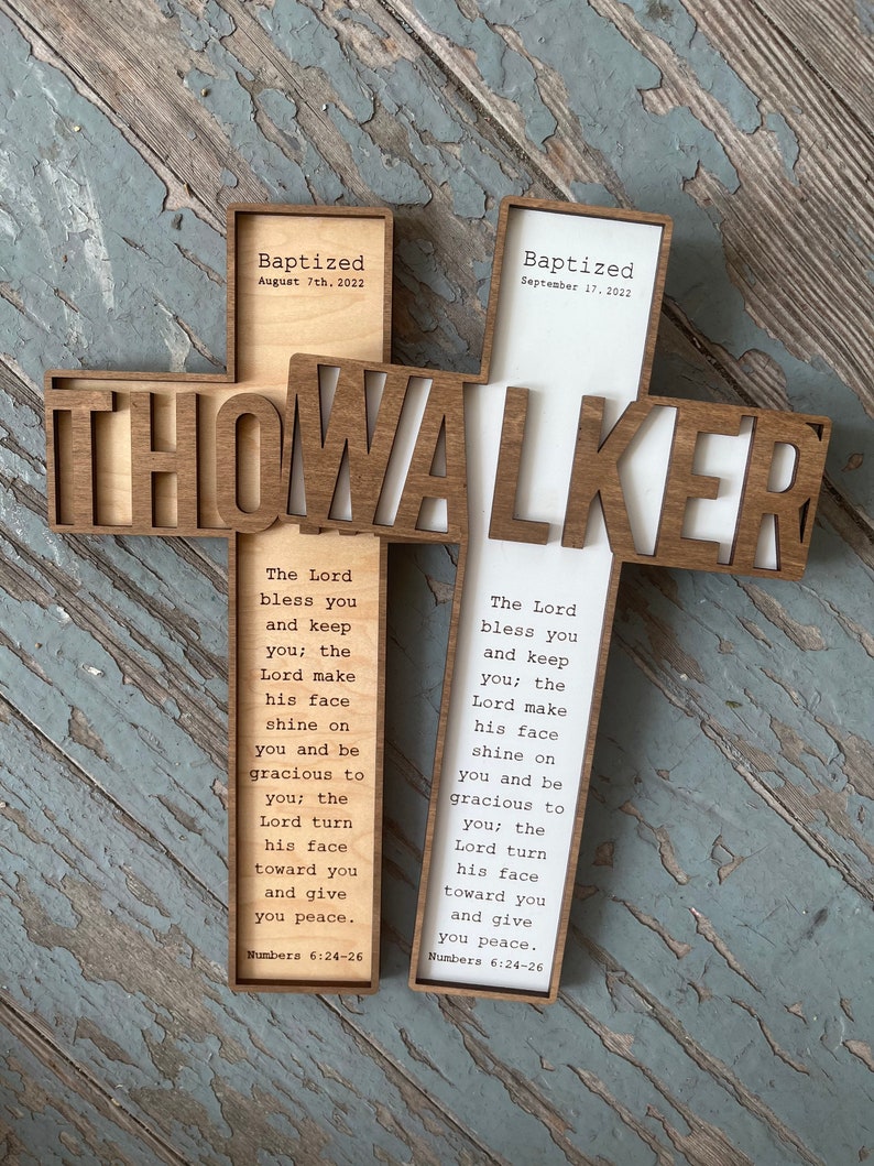 Peut inclure: Une croix en bois avec le nom "THOWALKER" d&eacute;coup&eacute; au centre. La croix est divis&eacute;e en deux sections, chacune avec une date de bapt&ecirc;me diff&eacute;rente et un verset biblique de Nombres 6:24-26. Le verset dit : "Que l'&Eacute;ternel te b&eacute;nisse et te garde ; que l'&Eacute;ternel fasse briller sa face sur toi, et qu'il te fasse gr&acirc;ce ; que l'&Eacute;ternel tourne sa face vers toi, et qu'il te donne la paix."
