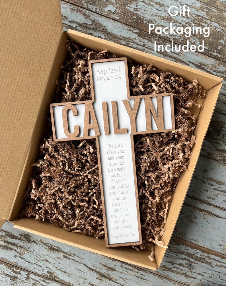 Peut inclure: Une croix en bois avec le nom "CAILYN" grav&eacute; dessus. La croix est dans une bo&icirc;te brune avec du papier d&eacute;chiquet&eacute;. La croix porte &eacute;galement le texte "Baptis&eacute; le 11 mai 2024" et un verset biblique : "L'&Eacute;ternel te b&eacute;nisse et te garde : l'&Eacute;ternel fasse briller sa face sur toi et te fasse gr&acirc;ce : l'&Eacute;ternel tourne sa face vers toi et te donne la paix, Nombres 6:24-26"