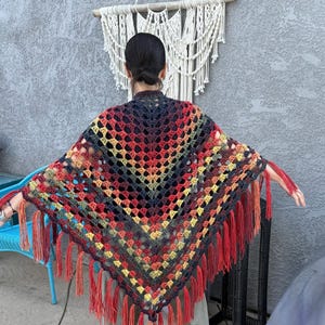 Puede incluir: Un chal de ganchillo hecho a mano y colorido con un patrón geométrico. El chal presenta un degradado de colores, incluyendo rojo, amarillo y azul, y está terminado con largas borlas rojas.