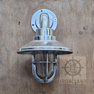 May include: A silver-colored wall-mounted light fixture with a clear glass shade and a protective cage. The fixture has a circular mounting plate and a curved top. The background is a wooden surface.