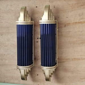 Pode incluir: Um par de arandelas de parede com painéis de vidro canelado azul-escuro dispostos verticalmente e emoldurados em metal dourado escovado. Cada luminária apresenta um design escalonado nas extremidades, criando uma silhueta cilíndrica alongada.