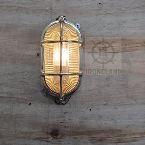 Op de afbeelding: Een messingkleurige, ovale wandlamp met een beschermende kooi en geribbeld glas. De lamp is verlicht en geeft een warme gloed. De armatuur is gemonteerd op een houten oppervlak. Het woord "IRONCLAND" is zichtbaar.