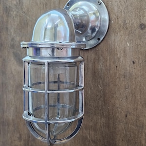 May include: A silver-colored, industrial-style wall-mounted light fixture. The fixture features a rounded top, a protective cage around the glass bulb, and a circular mounting plate. The light is designed to be mounted on a wall.