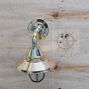 Może przedstawiać: Mosiężna lampa ścienna w stylu marynistycznym. Oprawa ma okrągłą podstawę, zakrzywione ramię i stożkowy klosz z ochronną klatką drucianą wok&oacute;ł szkła. Słowo "IRONCLAND" jest widoczne na drewnianym tle.