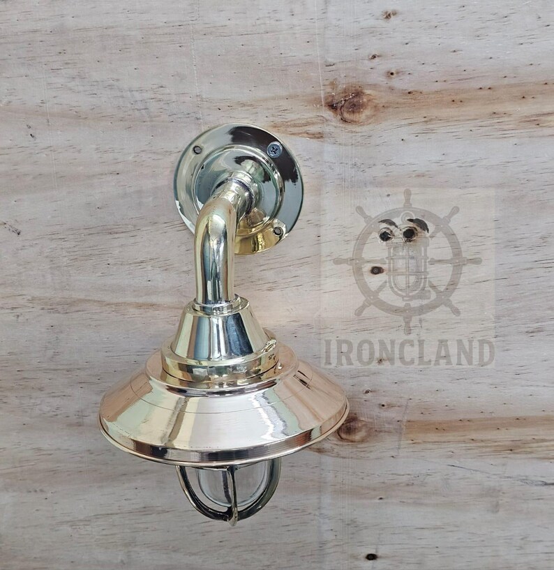 Może przedstawiać: Mosiężna lampa ścienna w stylu marynistycznym. Oprawa ma okrągłą podstawę, zakrzywione ramię i stożkowy klosz z ochronną klatką drucianą wok&oacute;ł szkła. Słowo "IRONCLAND" jest widoczne na drewnianym tle.