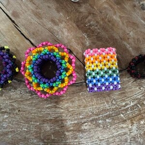Könnte beinhalten: Eine Auswahl an farbenfrohen Perlenaccessoires, darunter ein lila-schwarzes Perlenarmband, ein regenbogenförmiges Blumenelement, ein rechteckiges Regenbogenelement und ein schwarz-rotes Perlenarmband.