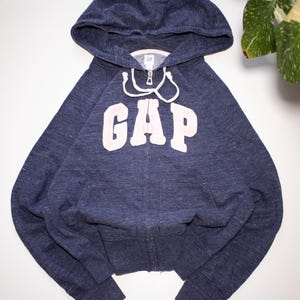 Peut inclure: Un sweat à capuche zippé bleu marine chiné avec le mot GAP en grandes lettres blanches sur la poitrine. Le vêtement comprend une capuche avec des cordons de serrage blancs, des manches longues, des poignets côtelés et deux poches avant.