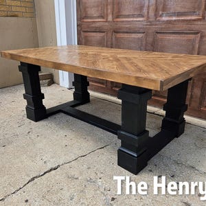 May include: A rectangular wooden table with a light brown tabletop and black legs. The tabletop has a patterned design. The legs are thick and blocky, with a sturdy, geometric design. The table is set on a concrete surface.