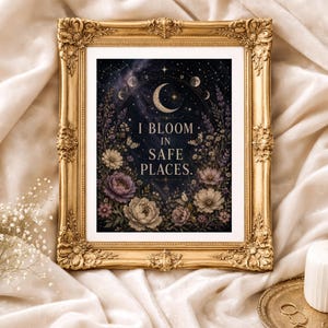 May include: A framed art print with a gold ornate frame. The artwork features a celestial design with a crescent moon, stars, and phases of the moon. The text "I BLOOM IN SAFE PLACES" is centered, surrounded by floral elements in shades of purple, cream, and green.