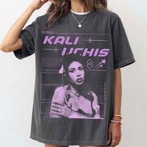May include: Dark gray t-shirt featuring a graphic of Kali Uchis in lavender. The design includes the artist's name, geometric shapes, and text. The shirt has a vintage, worn look.