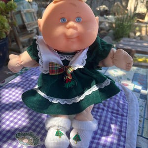 May include: A Cabbage Patch Kids doll in a green corduroy dress with white lace trim and a tartan bow. The doll has blue eyes and blonde hair. It is wearing white socks with a Christmas tree design.