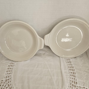 May include: Two off-white, oval-shaped ceramic dishes with handles. The dishes are empty and have a simple, classic design. They are placed side by side on a white, textured surface.