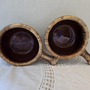 May include: Two brown ceramic mugs with handles. The mugs have a textured, speckled exterior with a lighter brown rim. The interior of the mugs is a darker brown. The mugs are sitting on a white, lace-trimmed surface.