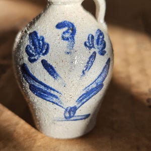 May include: Small, off-white ceramic jug with a handle and blue floral designs. The jug has a speckled texture and a rounded body. The blue design includes a central shape and two floral-like elements on either side. A charming decorative piece.