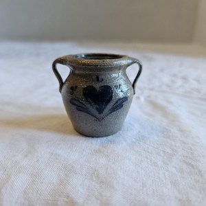 May include: Small, gray ceramic vase with two handles and a dark blue heart design. The vase has a rustic, handmade appearance, with a slightly rough texture. The heart is surrounded by stylized leaves.