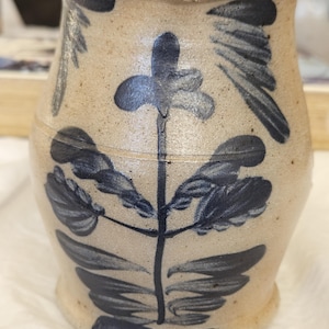 May include: A tan stoneware jar with a dark blue floral design. The jar has a cylindrical shape with a slightly flared rim and base. The floral design features a central stem with leaves and a flower at the top. The jar is likely handmade.