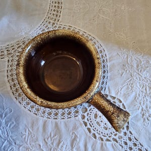 May include: A brown ceramic soup bowl with a handle. The bowl has a glossy finish and a speckled, textured rim. The interior is a darker brown, and the handle is a lighter brown with darker speckles.