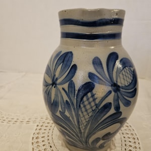 May include: A gray stoneware pitcher featuring a rounded body and a narrow neck with two horizontal blue bands. The surface is decorated with hand-painted cobalt blue floral motifs and cross-hatched patterns encircling the exterior of the vessel.