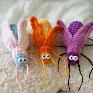 Puede incluir: Tres juguetes de insectos hechos a mano con ganchillo en rosa, naranja y morado. Cada insecto tiene grandes ojos saltones y largas antenas. Los juguetes están hechos de hilo suave y texturizado y descansan sobre una superficie blanca y esponjosa.