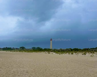 May include: A coastal scene featuring a sandy beach under a dramatic, cloudy sky. A tall, white lighthouse with a red top stands in the distance, framed by green vegetation. The image has a natural, serene quality.