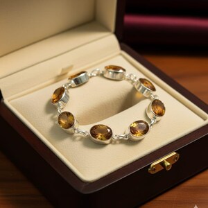 以下が含まれることがあります： 木製のジュエリーボックスの中に、楕円形、琥珀色の宝石が付いたシルバーブレスレットが展示されています。 ブレスレットはクリーム色のクッションの中央に配置され、ジュエリーを明らかにするためにボックスが開いています。