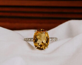 May include: A gold ring featuring a large, oval-cut citrine gemstone. The band is adorned with small, clear accent stones. The ring is set against a white fabric background, with a dark wooden backdrop.
