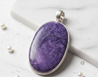 May include: Oval-shaped pendant with a polished, deep purple stone set in a silver-colored metal frame. The stone has a unique, swirled pattern with dark veins. The pendant is displayed with scattered pearls and sparkles.