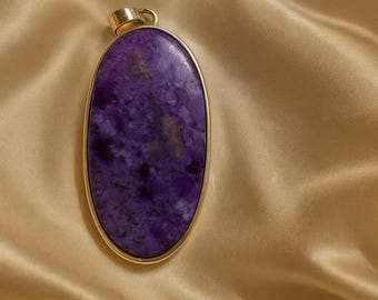 May include: Oval pendant featuring a large, polished purple stone with a gold-colored metal frame. The stone has a marbled appearance with shades of lavender and darker purple. The pendant has a small loop at the top for a chain.