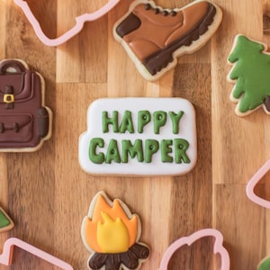 May include: A collection of camping-themed cookies and cookie cutters on a wooden surface. The cookies include a backpack, boot, campfire, mountains, and a sign that says "HAPPY CAMPER". Pink plastic cookie cutters are also present.