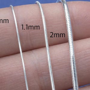 May include: Close-up of three silver snake chains in varying thicknesses: 0.8mm, 1.1mm, and 2mm. The chains are displayed against a skin-toned background, showcasing their texture and shine. The image highlights the different chain sizes.