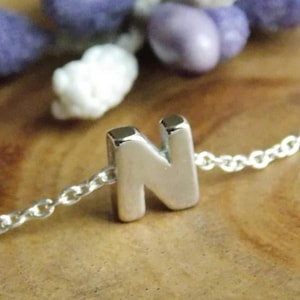 May include: A silver necklace featuring the letter "N" pendant. The chain is fine, and the pendant is centrally placed. The background includes purple and white decorative items on a wooden surface. This necklace is a personalized jewellery item.