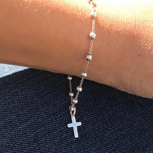 May include: Silver rosary bracelet with small, round beads and a cross pendant. The bracelet is worn on a wrist, with the focus on the delicate chain and religious symbol. The bracelet is a religious accessory.
