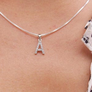 May include: A silver necklace with a pendant in the shape of the letter "A". The pendant is encrusted with small, clear gemstones. The necklace is worn against a backdrop of a white top with black heart-shaped patterns.