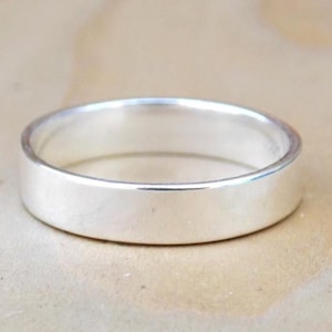 May include: A plain, polished silver ring. The band is flat and wide, with a smooth, reflective surface. The ring is a classic design, suitable for everyday wear or special occasions.