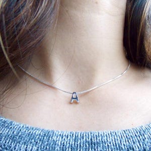 Peut inclure: Un collier en argent avec un petit pendentif en forme de lettre "A" majuscule. Le collier est porté sur un pull bleu. La chaîne est fine et le pendentif est centré, créant un look simple et élégant. Le collier est un bijou.