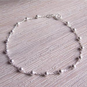 May include: A delicate silver bracelet with small, round beads spaced along a fine chain. The bracelet has a simple clasp. The jewellery is displayed on a light-coloured, textured surface.