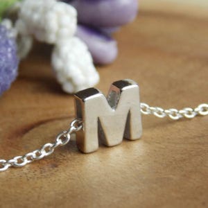 May include: A silver necklace with the letter "M" as a pendant. The pendant is a three-dimensional, block-style letter. The chain is a delicate, linked design. The background is a wooden surface with blurred purple and white decorative elements.