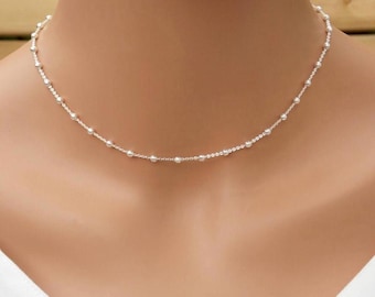 May include: A delicate silver necklace featuring small, evenly spaced beads. The necklace is displayed on a person's neck, showcasing its simple yet elegant design. The beads appear to be a bright, reflective silver.