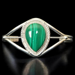 May include: Silver cuff bracelet with a striking, teardrop-shaped malachite stone in vivid green. The stone is framed by a textured border. The bracelet has a geometric design and a polished finish, set against a black backdrop.