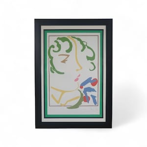 May include: Framed art print featuring a stylized profile of a person with green hair, yellow accents, and a pink lip. The artwork includes blue and red floral elements. The print is set within a green and brown border, encased in a black frame.