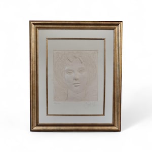 May include: A framed artwork featuring a bas-relief portrait of a woman with curly hair. The artwork is in a gold-colored frame with a white mat. The portrait is in a light color, possibly plaster or a similar material.