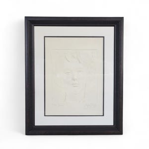May include: A framed artwork featuring a detailed, embossed portrait of a person's face. The artwork is in a white color, set against a white mat, and enclosed in a dark brown, ornate frame. The image is centered on a white background.